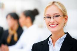 Blonde office woman with glasses smiling with office co workers in background