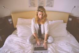 Blonde woman sitting on her bed whilst working on her laptop