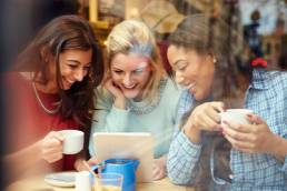 Three women smiling, procrastiniating,using digital devices whilst drinking in a cafe