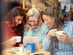 Three women smiling, procrastiniating,using digital devices whilst drinking in a cafe