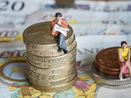 Man sitting on big pile of coins next to woman sitting on smaller pile of coins to demonstrate gender gap