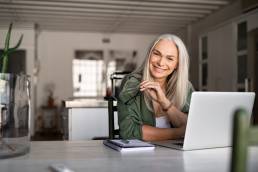 Portrait of happy senior woman holding eyeglasses and looking at camera at home. Successful old lady laughing and working at home, menopause