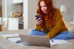 Attractive Young Woman with Red Hair Working from Home - Female Entrepreneur Sitting on Bed with Laptop Computer, Paperwork and Checking Cell Phone from Comfort of Home