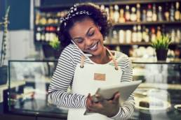 Smiling young African entrepreneur standing in front of the counter of her cafe talking on a cellphone and using a tablet, single parent business owner