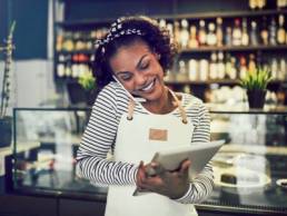 Smiling young African entrepreneur standing in front of the counter of her cafe talking on a cellphone and using a tablet, single parent business owner