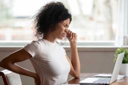 Tired african American millennial female worker sit at desk touch back suffer from lower spinal spasm, working from home