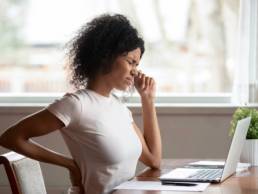 Tired african American millennial female worker sit at desk touch back suffer from lower spinal spasm, working from home