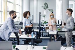 Modern team meeting, group work and social distancing. Manager with tablet speaks with workers in protective masks in interior of modern office with gadgets during coronavirus epidemic, free space