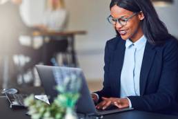 Smiling young African American businesswoman working on a laptop at her desk in a bright modern office with colleagues in the background