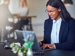 Smiling young African American businesswoman working on a laptop at her desk in a bright modern office with colleagues in the background