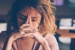 Closeup of sad young Asian woman at cafe leaning head on clasped hands and staring into vacancy. Tired freelancer feeling burnout. Stress and bad news concept