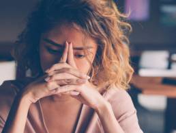 Closeup of sad young Asian woman at cafe leaning head on clasped hands and staring into vacancy. Tired freelancer feeling burnout. Stress and bad news concept