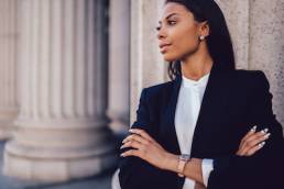 Female African American banker dressed in elegant black suit folding hands and looking on side standing against office building.