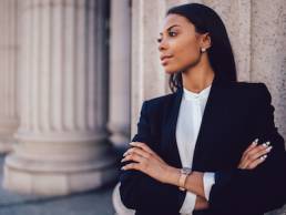 Female African American banker dressed in elegant black suit folding hands and looking on side standing against office building.