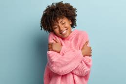 Love yourself concept. Photo of lovely smiling woman embraces herself, has high self esteem, closes eyes from enjoyment, likes her new comfortable soft pink sweater, tilts head, stands indoor. self care