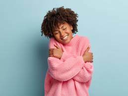 Love yourself concept. Photo of lovely smiling woman embraces herself, has high self esteem, closes eyes from enjoyment, likes her new comfortable soft pink sweater, tilts head, stands indoor. self care