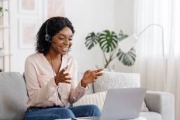 Video Conference. Smiling african woman having web call on laptop at home, talking at camera while sitting on sofa in living room, flexible working