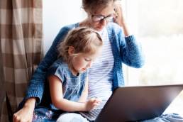 Working mom works from home office with kid. Mother and daughter read news. Woman and cute child sitting on window sill.