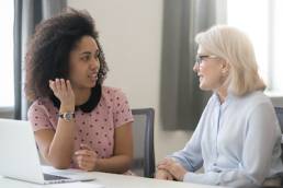 Diverse old and young female colleagues talking at work, african and caucasian business women sitting together in office having friendly conversation, mentor intern discussing planning shared project, Menopause