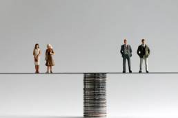 A group of miniature men and women standing on either side of a pile of coins, equal pay, gender pay gap