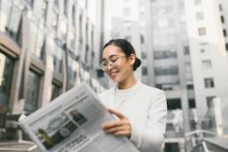 Young attractive Asian female banker or accountant in glasses is reading newspaper outside a modern office center or a bank