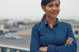 Confident friendly black business woman standing with folded arms on the rooftop of an urban commercial building smiling as she looks to the side of the camera, self-acceptance