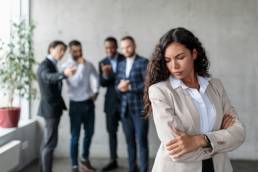 Workplace Sexism And Bullying. Unhappy Victimized Businesswoman Standing While Her Male Colleagues Whispering Behind Her Back Standing In Modern Office