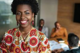 close up of beautiful young smiling professional black african business woman, coworkers hold a meeting in background, black female founders list