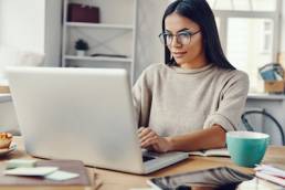 Beautiful young woman in casual clothing using laptop and smiling while working indoors