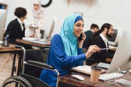 Disabled arab woman in hijab in wheelchair working in office. Woman is working on desktop computer and talking on phone.