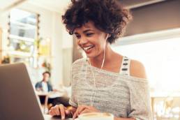 Image of happy woman using laptop while sitting at cafe. Young african american woman sitting in a coffee shop and working on laptop, workplace wellbeing