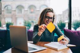 Positive hipster girl in stylish spectacles for better view using stickers for productive learning information from book, World Productivity Day