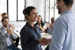 Happy proud excited Indian ethnicity employee get promotion receive praises from boss and cheering and congratulations from diverse staff members shake hands with chief. Recognition of success concept