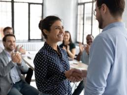 Happy proud excited Indian ethnicity employee get promotion receive praises from boss and cheering and congratulations from diverse staff members shake hands with chief. Recognition of success concept