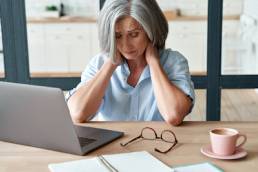 Tired stressed old mature business woman suffering from neckpain working from home office sitting at table. Overworked senior middle aged lady massaging neck feeling hurt pain from incorrect posture. Menopause