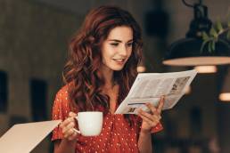 portrait of woman with cup of coffee reading newspaper at table with laptop in cafe