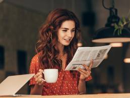 portrait of woman with cup of coffee reading newspaper at table with laptop in cafe