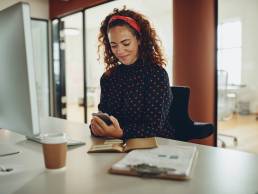 Young businesswoman smiling while reading text message on mobile phone sitting at desk in office