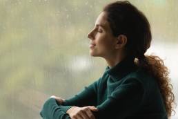 Side shot of serene woman relaxed by window with closed eyes listen sound of raindrops feel pleasure. Calm young lady enjoy breathing fresh ozonized air after rainstorm.