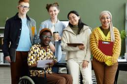 Portrait of diverse creative team looking at camera with cheerful smiles while posing in office, wheelchair user inclusion