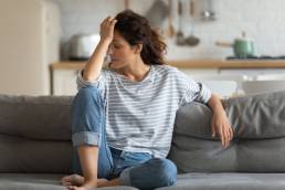 Exhausted frustrated young woman touching forehead, sitting on couch alone, suffering from strong headache, hormones or migraine, worried girl thinking about problems, divorce or break up, lost in thoughts